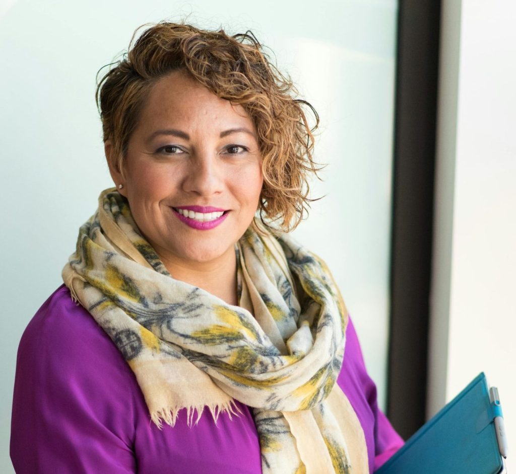 Smiling woman in a purple dress and scarf holding a tablet indoors.