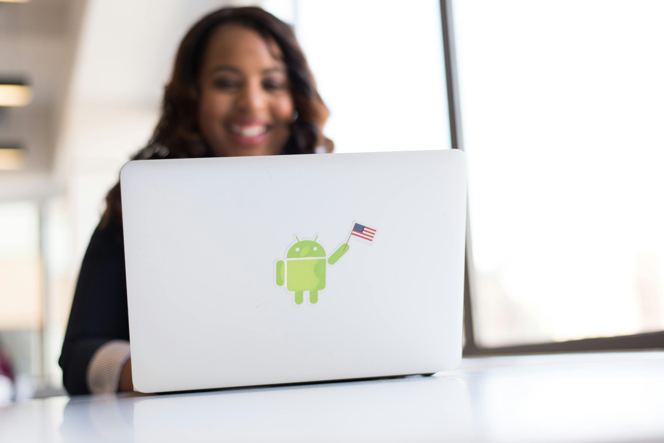 An African American woman working remotely on a laptop featuring an Android sticker in a bright office.